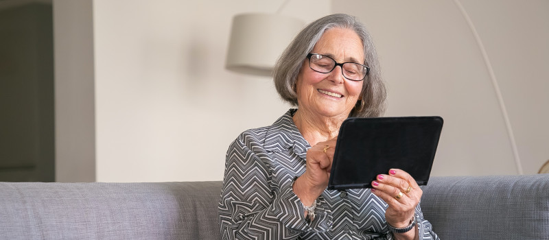 Mulher idosa a usar o novo serviço de Apoio Domiciliário com um tablet na mão e sorridente.